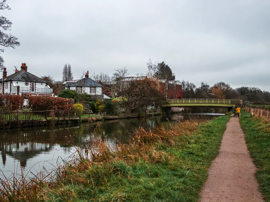 A trail next to the river, leading towards a bridge on the walk from Guildford to Godalming. There are some houses and their front lawns on the other side of the river.