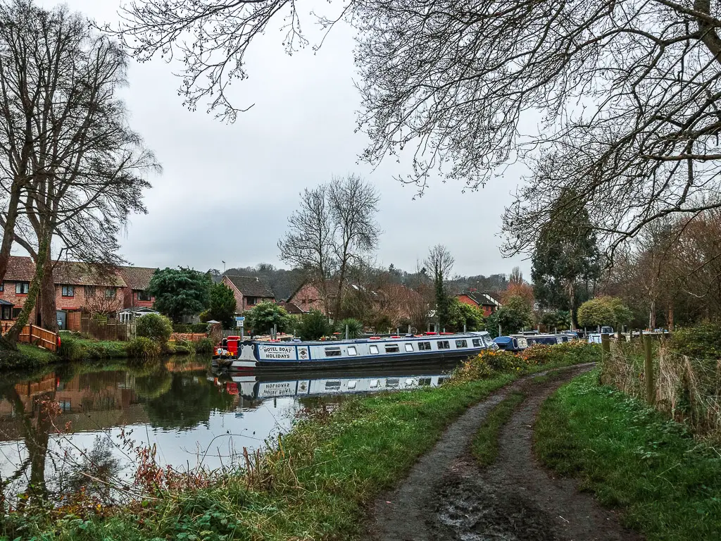 The dirt trail running along to the right, alongside the River Wey, on the walk into Godalming. there are some Barge boats on the river, and houses on the other side.