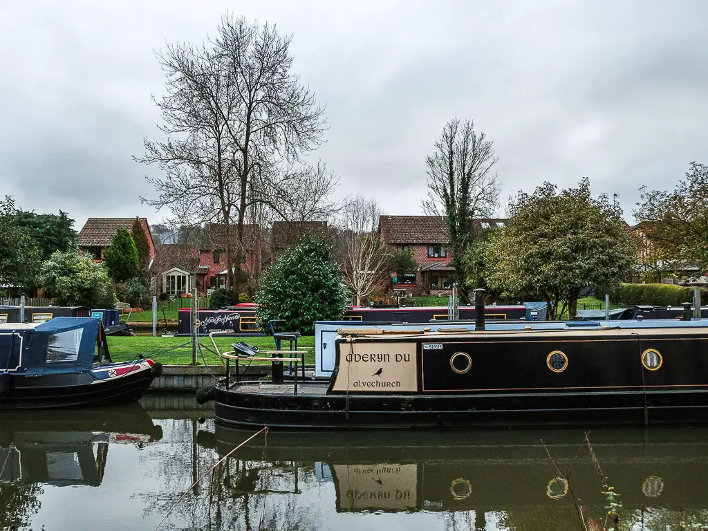 Barge boats moored along the River near Godalming, with houses and and dome trees on the other side.