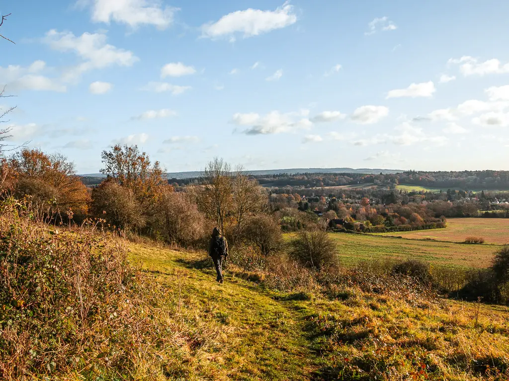 A man walking downhill with a view to the fields ands trees below in the distance.