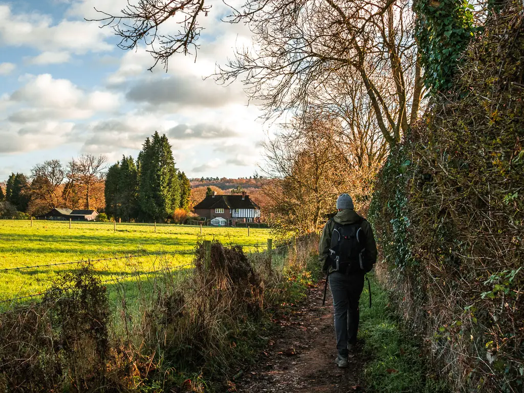 A man walking along a dirt track, with a hedge to the right and barbed wire fence and field to the left on the Newlands Corner St Martha's Hill walk. There is a house visible ahead.