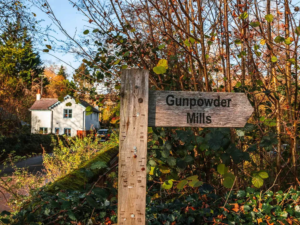 A wooden signpost saying Gunpowder mills in front of some greenery. There is a white coloured house visible to the left.