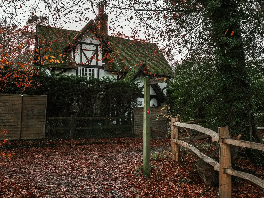 A period house behind a fence and bushes. The ground in front is covered in red fallen leaves and there is a wooden trail signpost.