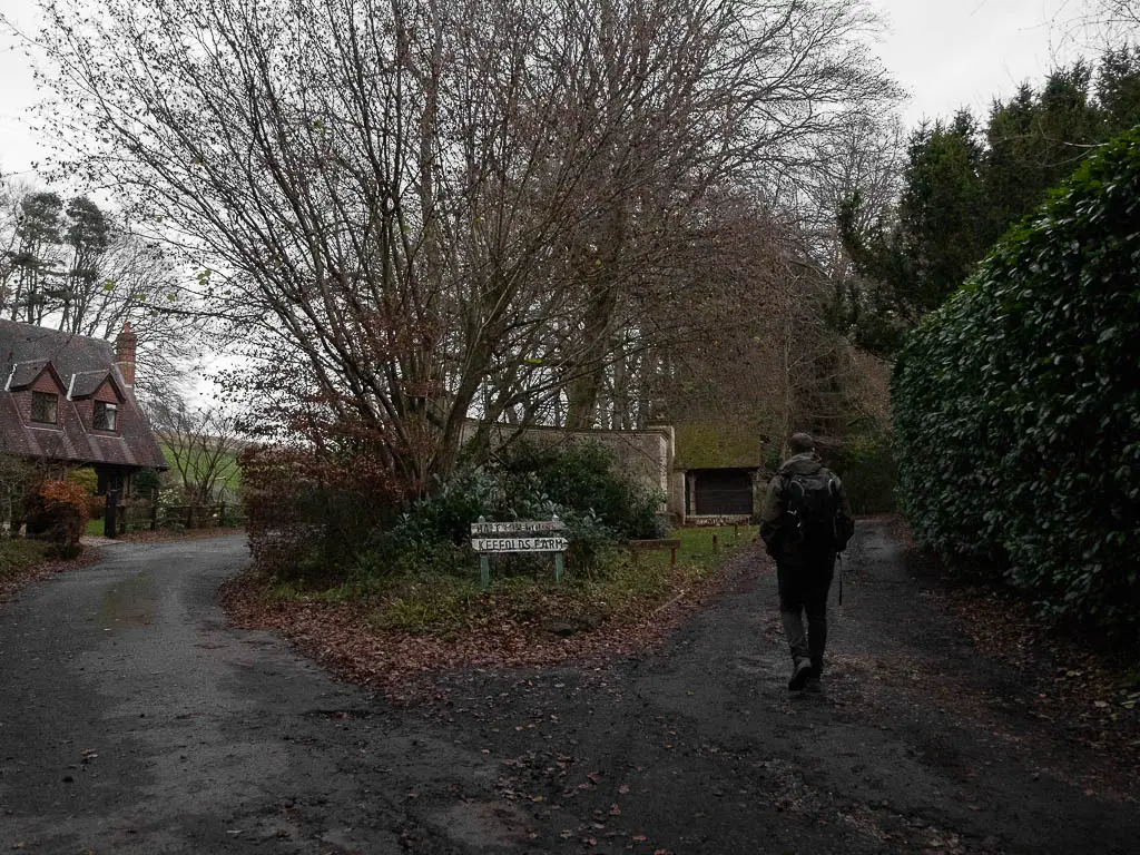 A fork in the road, with a hedge to the right, a house to the left and a road sign in the middle. There is a man walking up the right road.