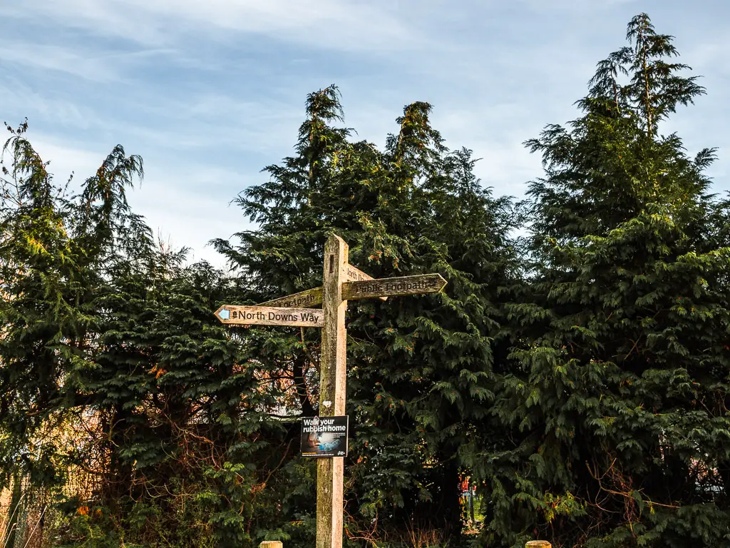 A wooden signpost marking the walk route for the North Downs Way stage 2. The signpost is in front of a group of leafy green trees.