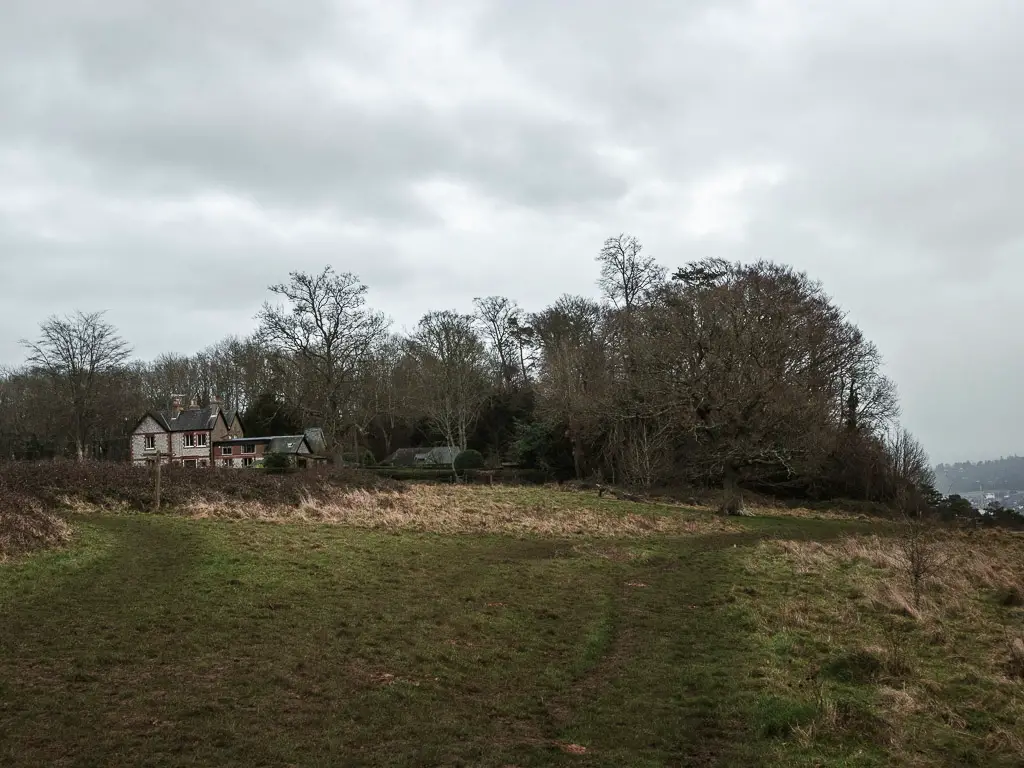 A large grass trail with woodland ahead and a cute house on the left behind a hedge on the walk from Guildford to Westhumble.