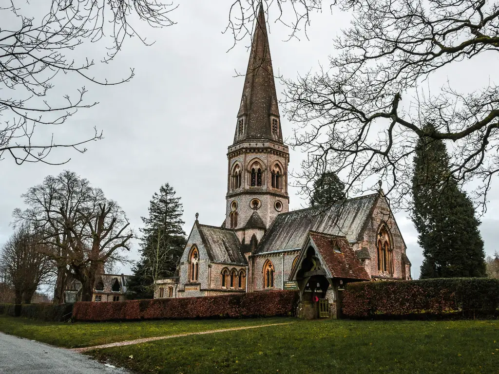 st Barnabas Church behind a hedge and surrounded by a few trees towards the end of the walk from Guildford to Westhumble. there are some leafless tree branches in the frame of the photo. 
