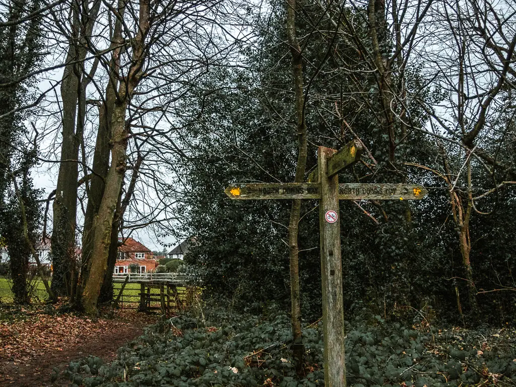 A wooden trail signpost at the end of the walk from Guildford to Westhumble. Ahead past the sign is a wooden gate leading into a field with some houses on the other side. 