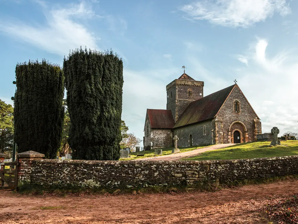 St Martha's Church, with a few gravestones visible. there is a stone wall around the perimeter of the church and two large hedges standing to the side.