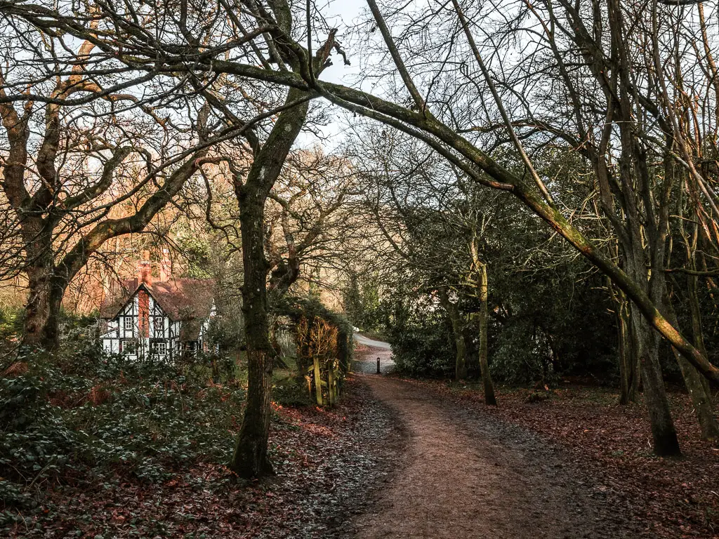 The dirt trail leading downhill through the trees towards a road. There is a black and white period house to the left through the trees.