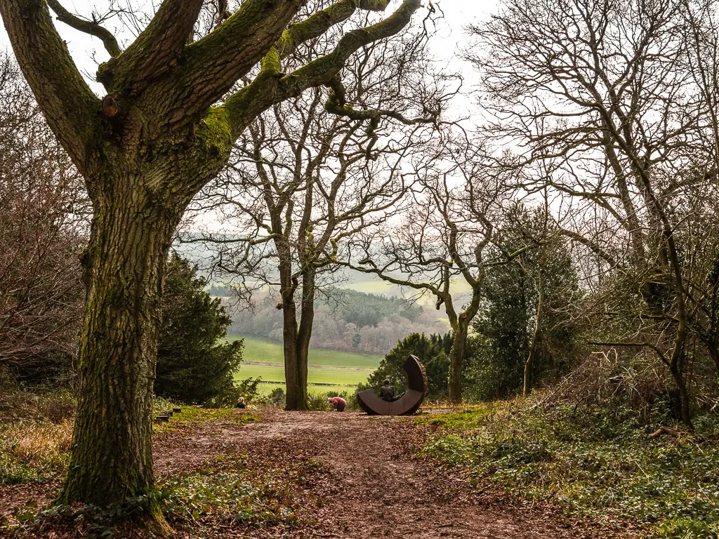 Looking through the trees to a C shaped art installation on the North Downs Way walk from Guildford to Westhumble. There is a view of the green fields through the trees.