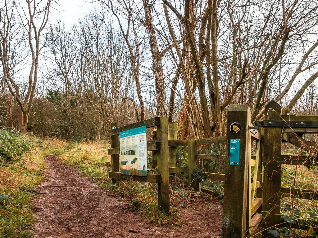 A mud trail running next to a wooden fence and gate. there is an information board on the gate and a black disc sign.