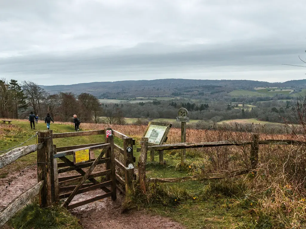Looking down to the wooden gate and fence leading into a hill field with green trees and hills in the distance on the walk from Guildford to Westhumble. there are a few people walking in the field. The sky is grey.