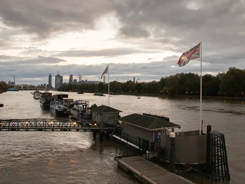 Looking down the River Thames along the pier at sunset. There is a Union Jack flag raised. On the other side of the river are the trees of Battersea Park.