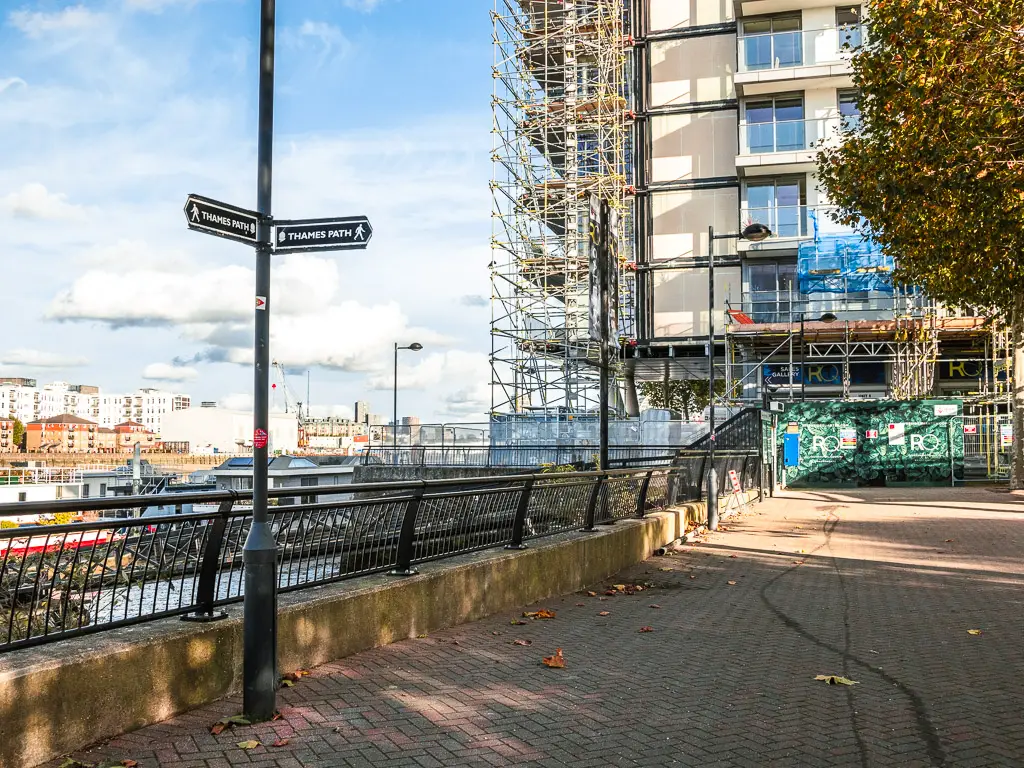 A black lamppost with Thames Path signage on the walk to Battersea from Putney. There is an apartment building behind with scaffolding up the side. 