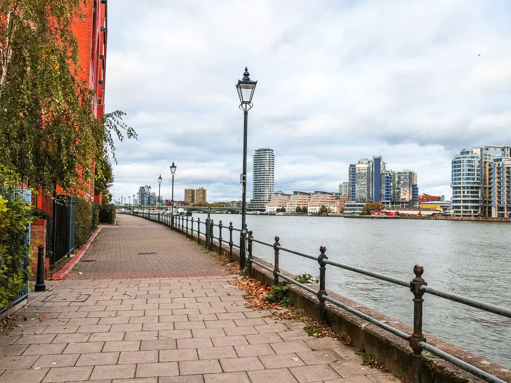 The Thames Path next to the river on the Putney to Battersea walk. There are tall apartment buildings on the other side of there river. 