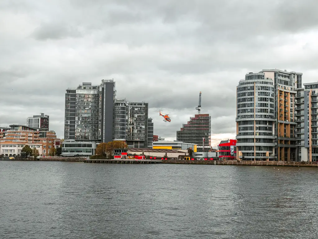 Looking across the river to the tall apartment buildings on the other side on the walk along the Thames Path from Putney to Battersea. there is a red helicopter about to land. 