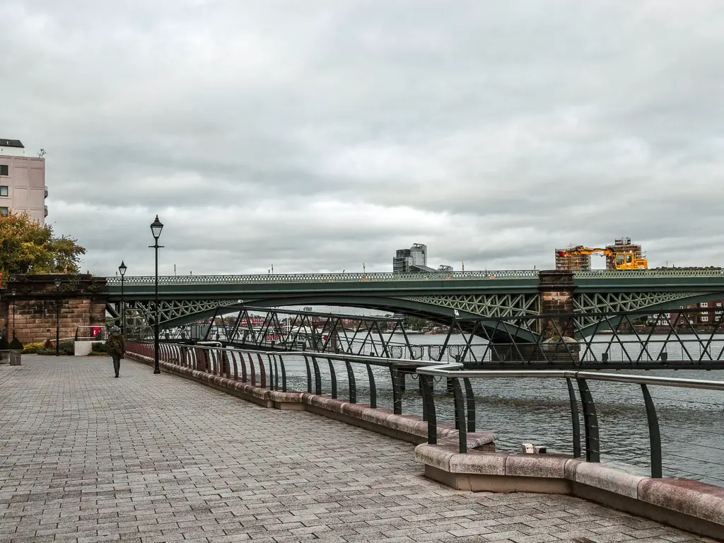 A man on a walk on the Thames Path on the route between Putney and Battersea. Ahead is a green railway bridge crossing the river. 