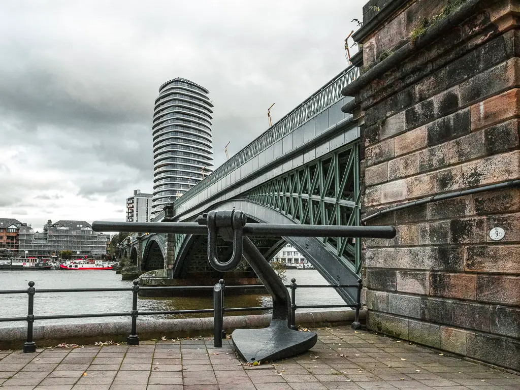 A giant metal anchor on the path next to a green bridge which runs across the River Thames. There is a tall apartment building on the other side of the bridge. 