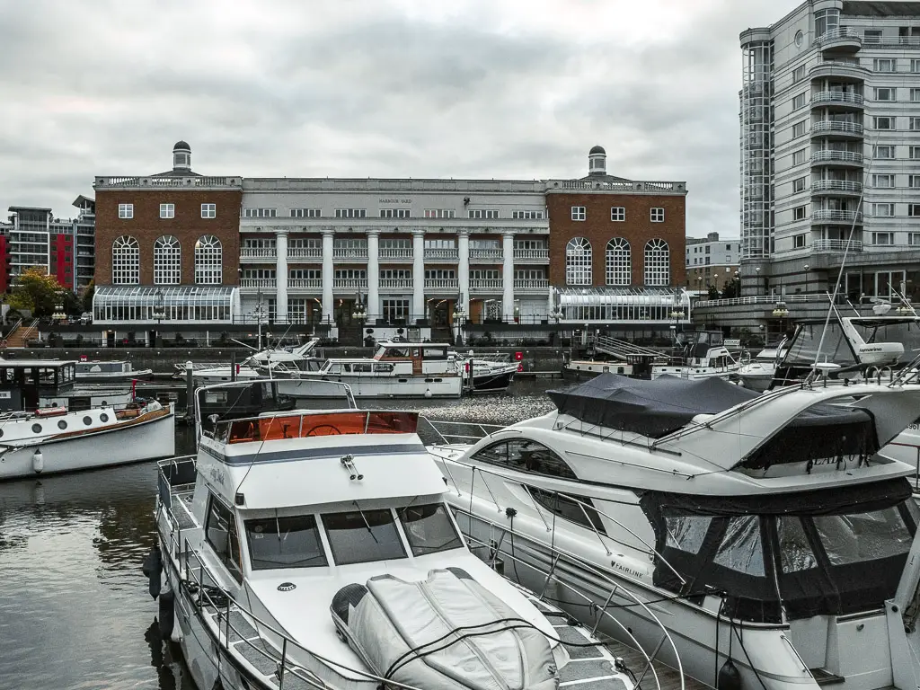 Lots of which yachts and boats docked in Chelsea Harbour. There is a large brown and white building on the other side. 