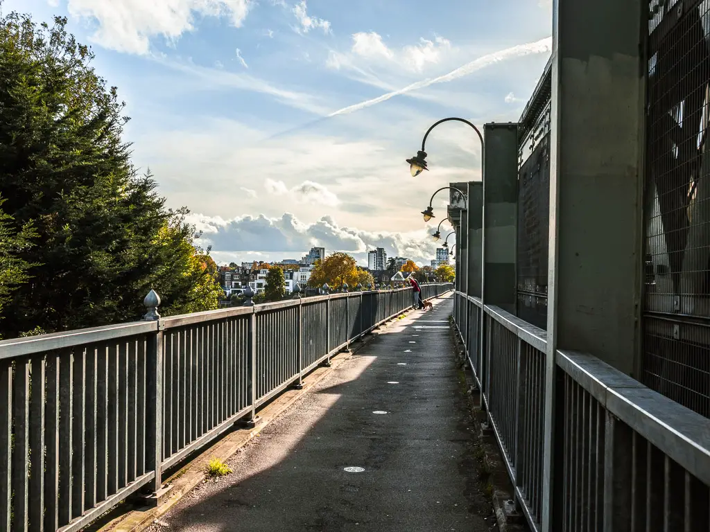 Looking along the footpath of Fulham Railway Bridge. There are a couple of people and their dog standing on the bridge looking over the railing. 