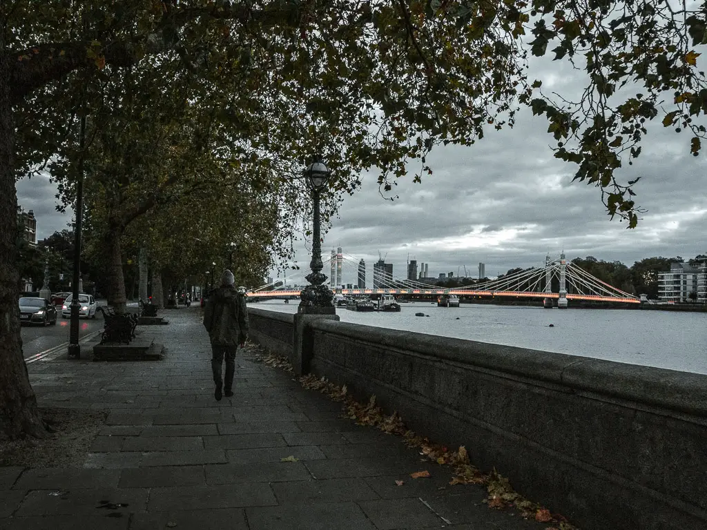 A man on a walk along the Thames Path on Chelsea Embankment towards the end of the walk from Putney to Battersea. The River Thames is on the right, and Albert Bridge, lit up, is further ahead. It is getting dark. 