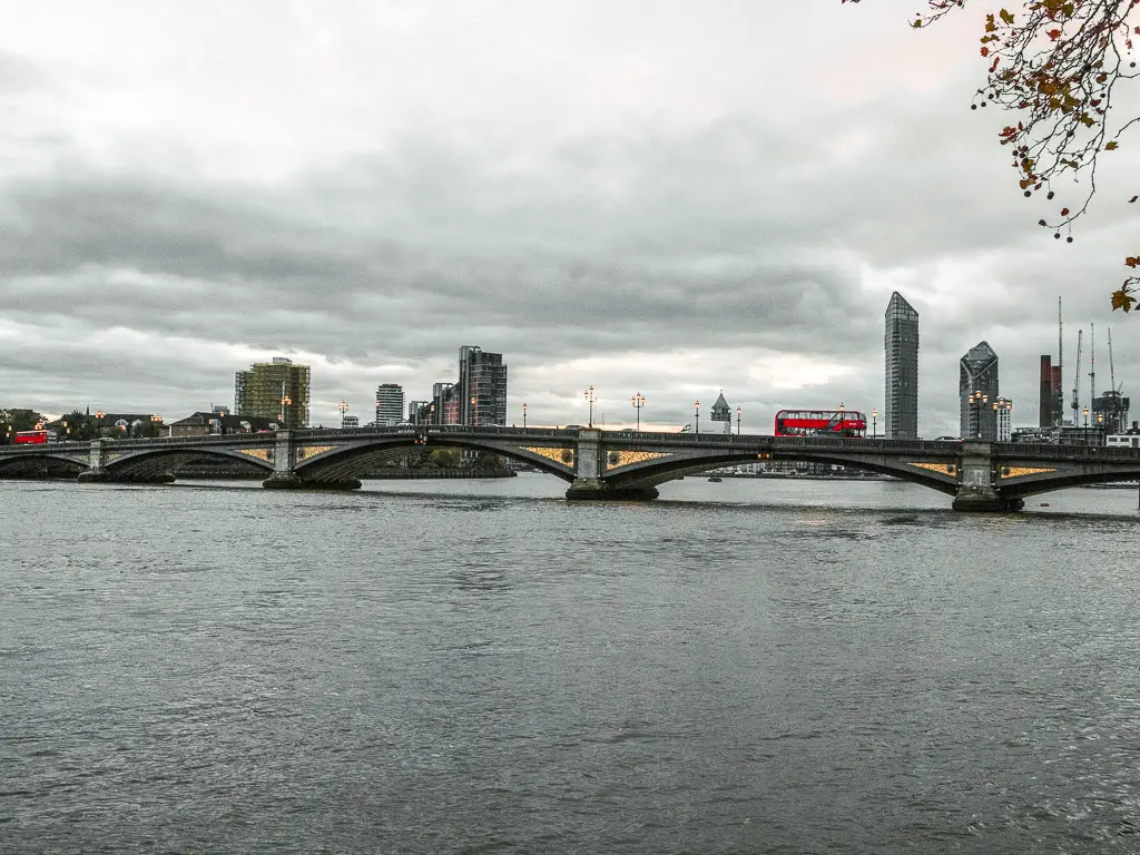 Looking along the River Thames to Battersea Bridge, with a red bus going over it. There are some tall apartment buildings visible in the diatcue on the other side. 