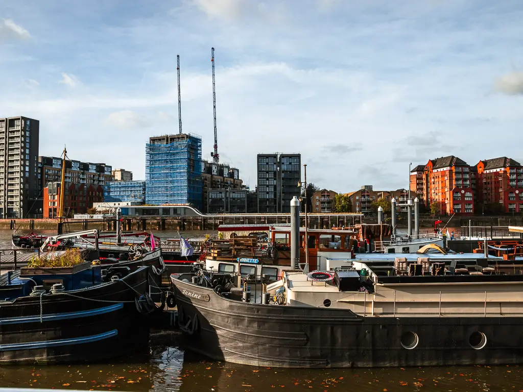 Barge boats moored on the river in Battersea on the walk along the Thames Path from Putney. There are red, blue and grey coloured apartment buildings on the other side. 