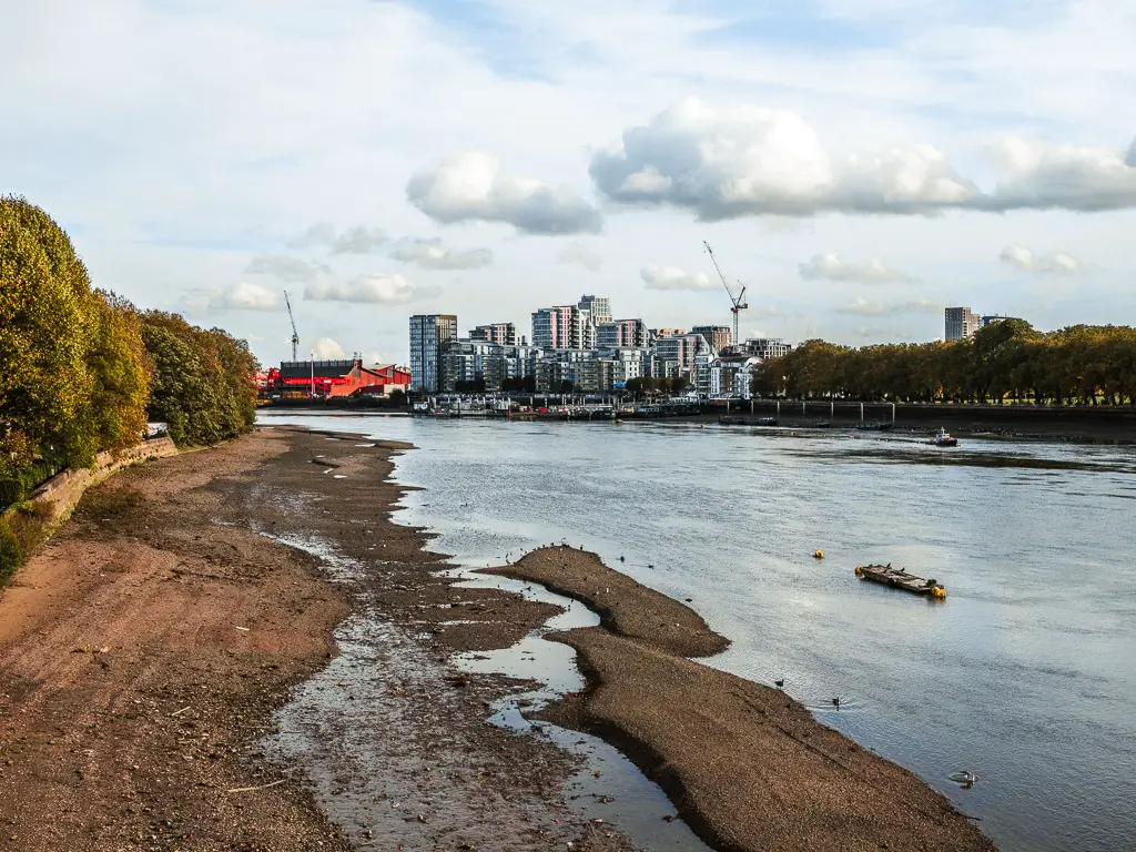 Looking down the bank of the River Thames at low tide on the Thames Path walk towards Battersea from Putney. there is a group of high rise apartments in the distance on the other side of the river. 