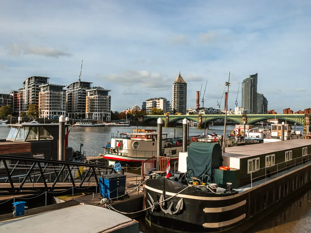 A group of barge boats moored on the side of the river on the walk from Putney to Battersea along the Thames Path. There is a green bridge going across the river, and apartment buildings on the other side.