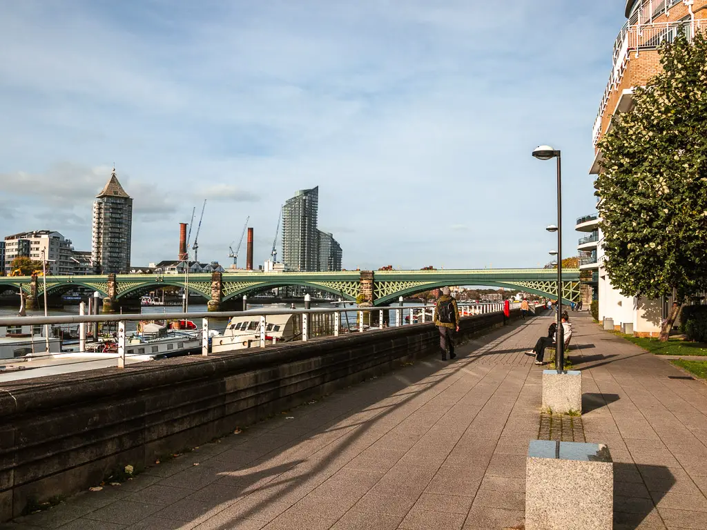 Looking along the Thames path on the Putney to Battersea walk. There are lamppost on the path, a woman sitting down on a bench and a man walking. There is a green coloured bridge ahead, crossing the river.