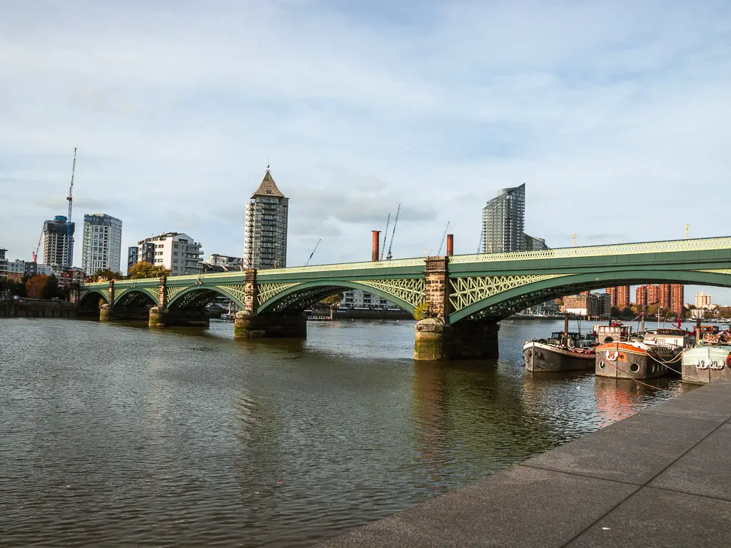 A green Galway bridge over the River Thames. There are a few barge boats moored under one of the arches of the bridge. Tall apartment buildings are visible in the distance. 