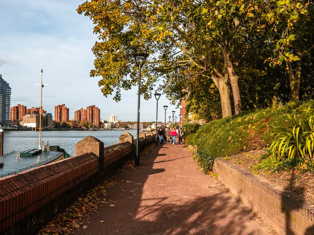 A group of people on a walk along the Thames Path in Battersea. The river is to the left and there are bushes and trees to the right.