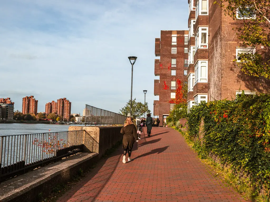 People walking along the path next to the River Thames on the walk from Putney to Battersea. There are apartment buildings on the right and ahead.