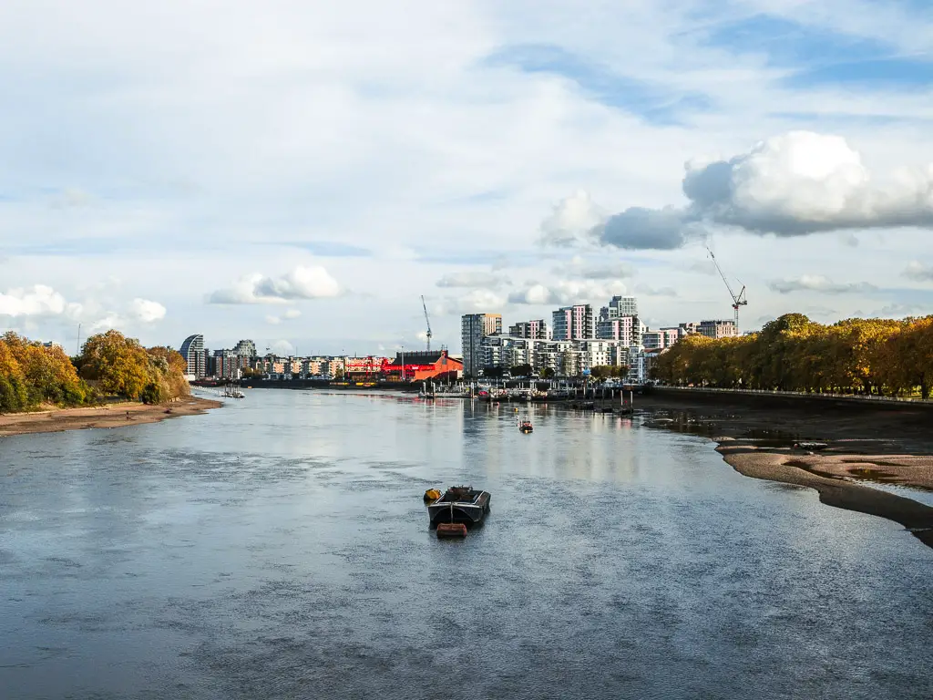 Looking across the River Thames, lined with trees and with high rise apartments in the distance. There is a small boat moored in the middle of the river. 