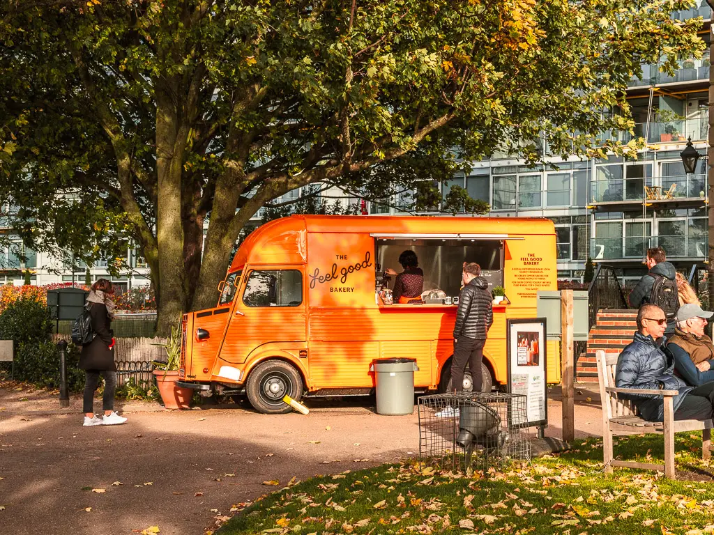 A bright orange food and coffee truck  in Battersea with a few people mingling about. There is a big tree behind the truck.