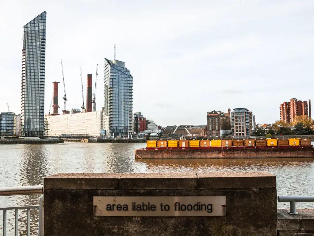 A sign saying 'area liable to flooding' infant of the railings of the River Thames. There is a cargo platform in the middle of the river and apartment buildings on the other side.