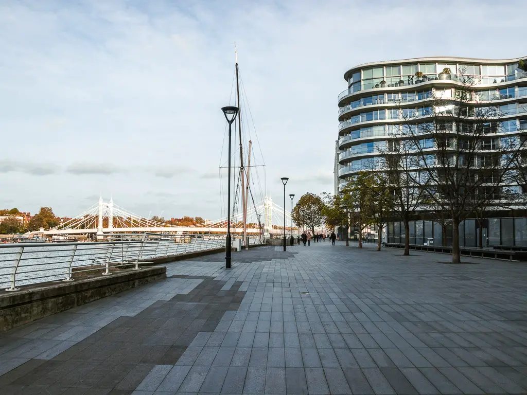 The large open Thames Path towards the end of the Putney to Battersea walk. There is a curved window covered apartment building to the right and the River Thames to the left. Albert Bridge is visible in the distance going across the river. 