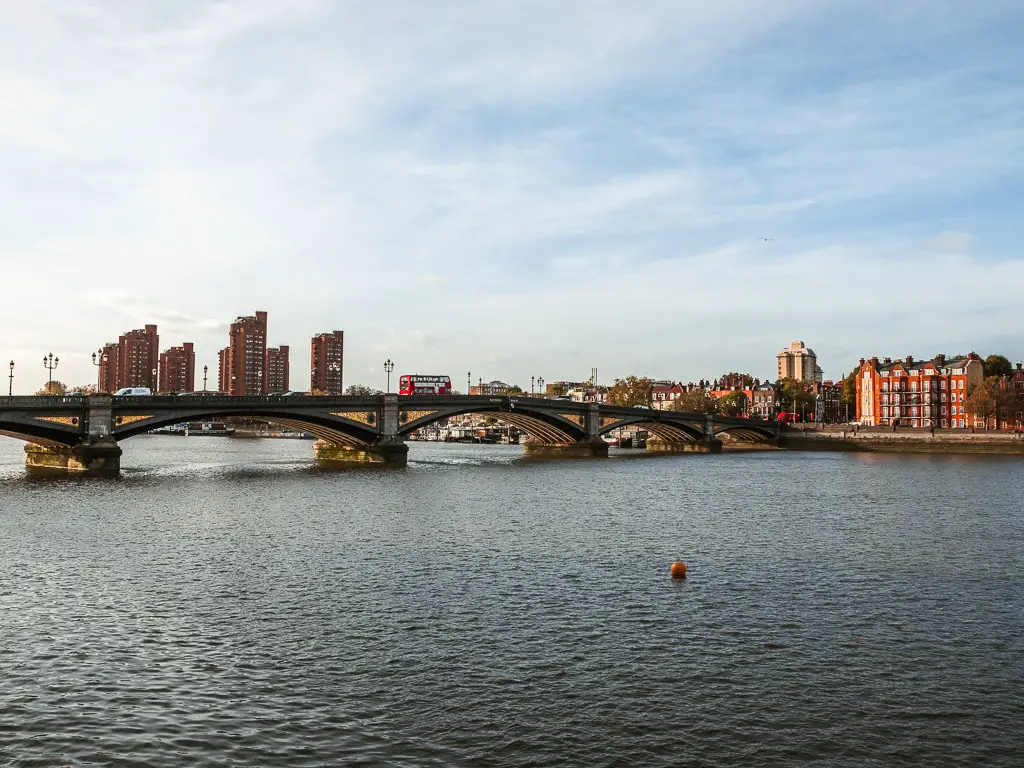 Looking across the River Thames to Battersea Bridge on the Thames Path walk from Putney. There is a red double decker bus going across the bridge, and brown high rise apartments in the distance on the other side. 