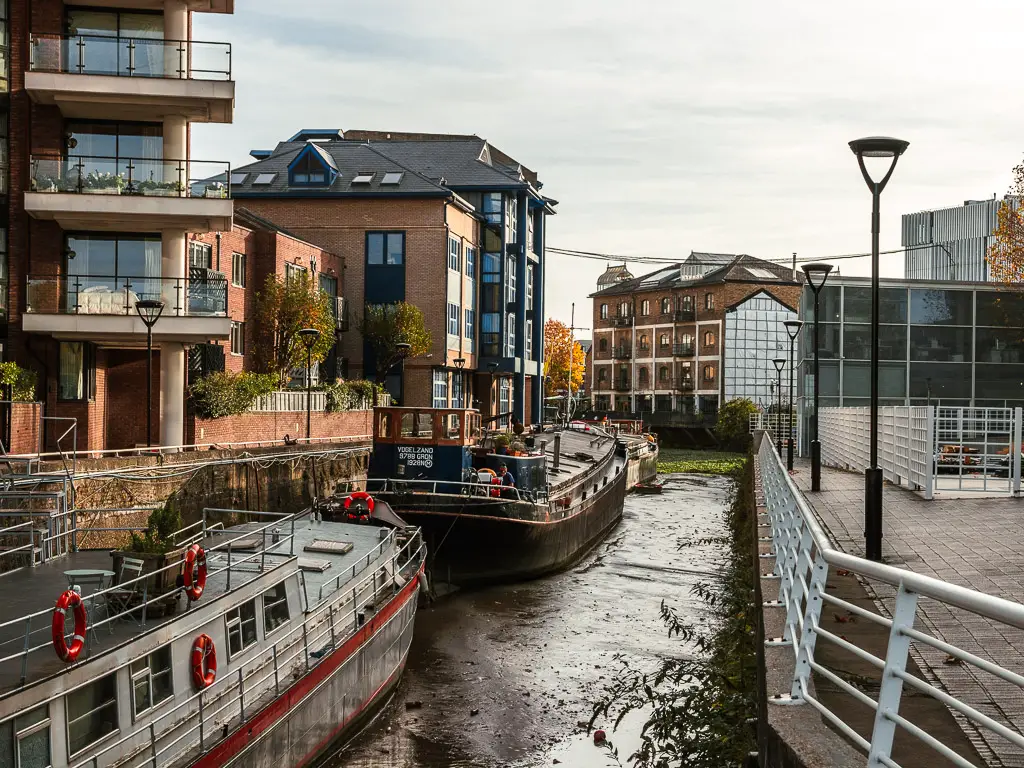 Looking along Ransom's Dock in Battersea. The tide is out, revealing the river bed. There are two barge boats next to the side, and apartments to the left. 