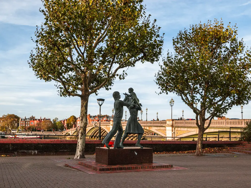 the 'In Town' statue on the Thames Path in Battersea on the walk from Putney. There are two trees next to the stature. Battersea Bridge is partially visible behind to the right. 