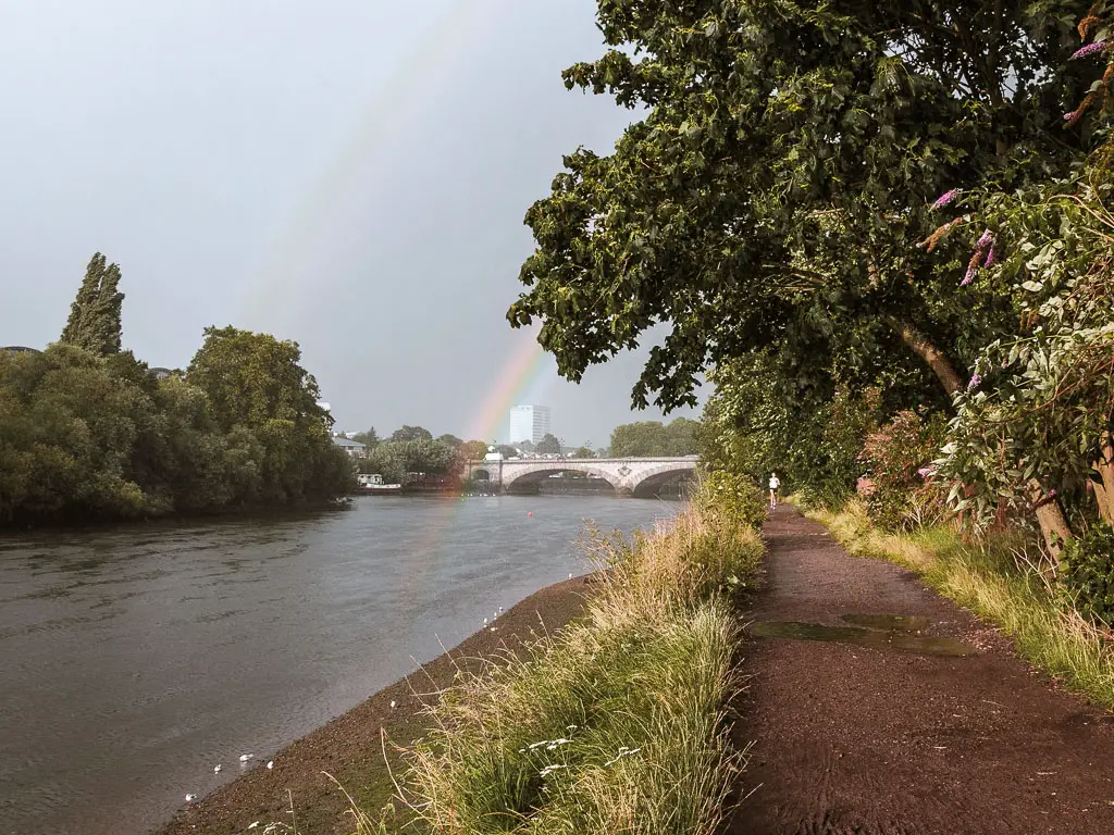 The Thames Path with trees to the right and the river to the eft on the Richmond to Putney walk. There is a rainbow coming out from the river, and a bridge further along in the distance. 