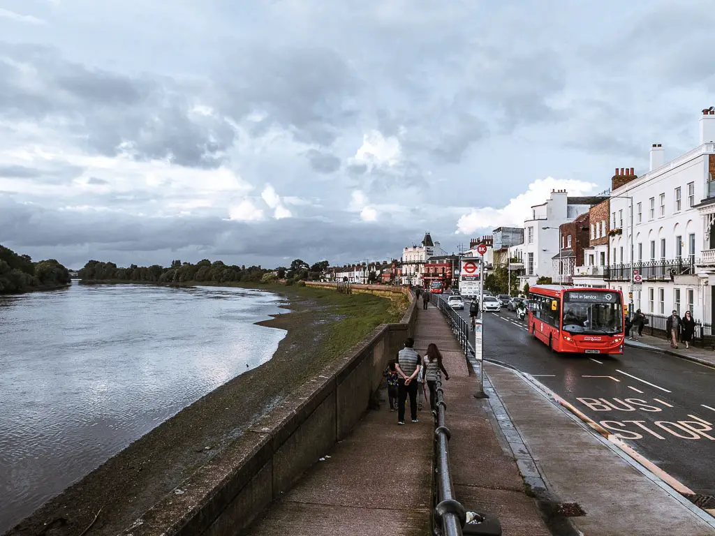 Looking down along the pavement which runs alongside the river which is to the left. To the right is the high street and a red bus driving on the road. There are a few people walking on the path. 