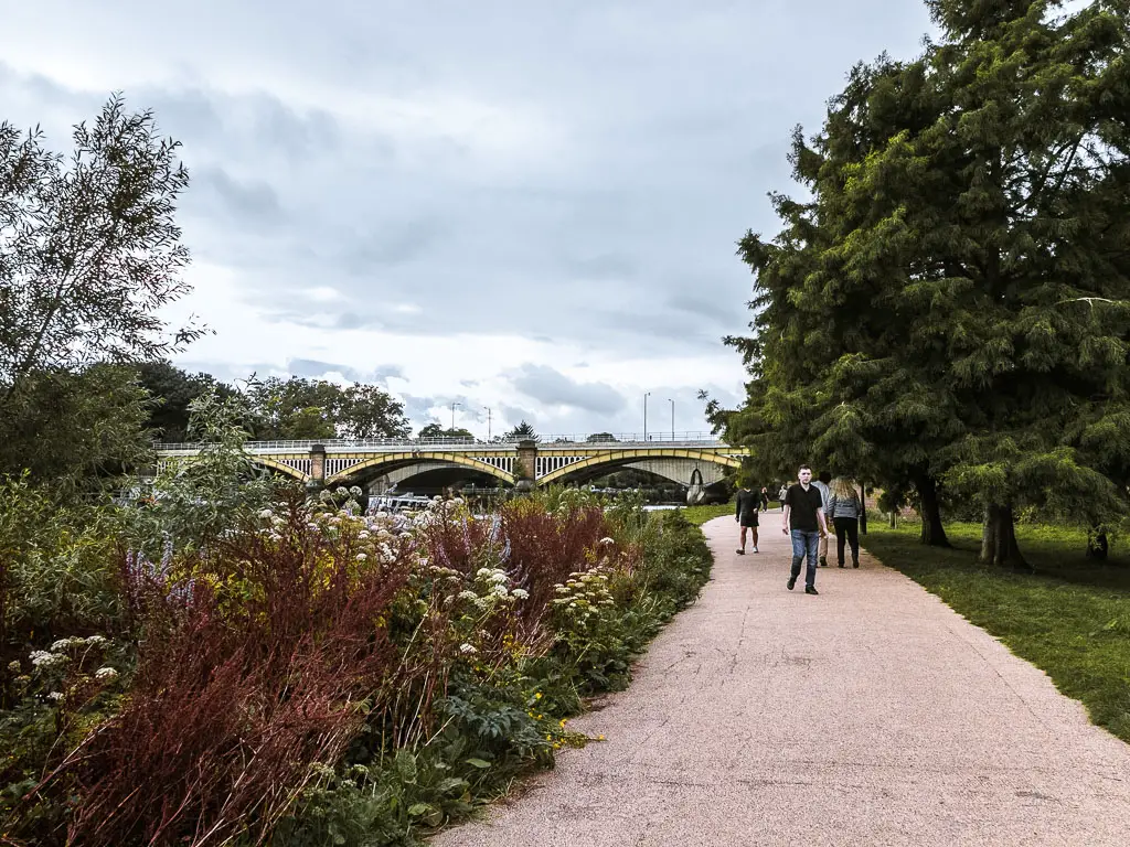 People walking along the Thames Path in Richmond. There are trees on the right, bushed on the left, and a bridge up ahead.