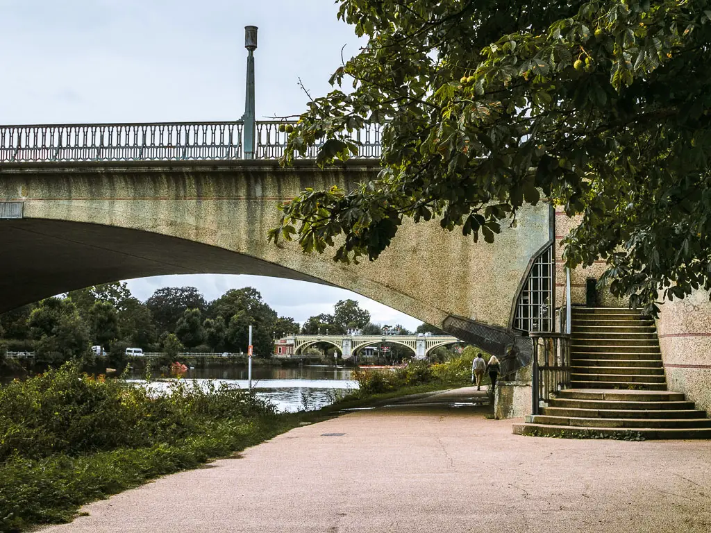 The Thames Path as it leads under a bridge. There is another bridge visible under and through the bridge. 