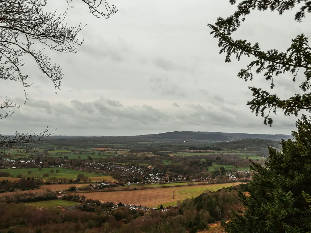 Looking through a gap in the trees to the view in the distance of fields and hills and a village when walking along stage 3 of the North Downs Way.