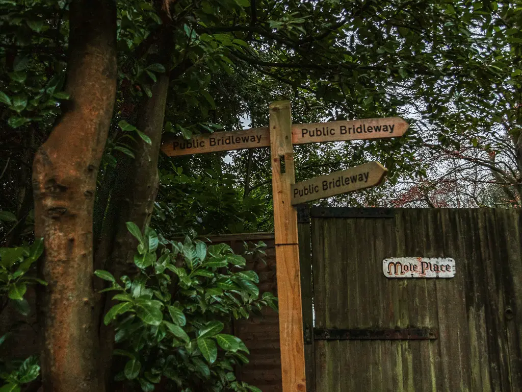 A wooden cross signpost standing in front of a wooden gate and next to a big tree.