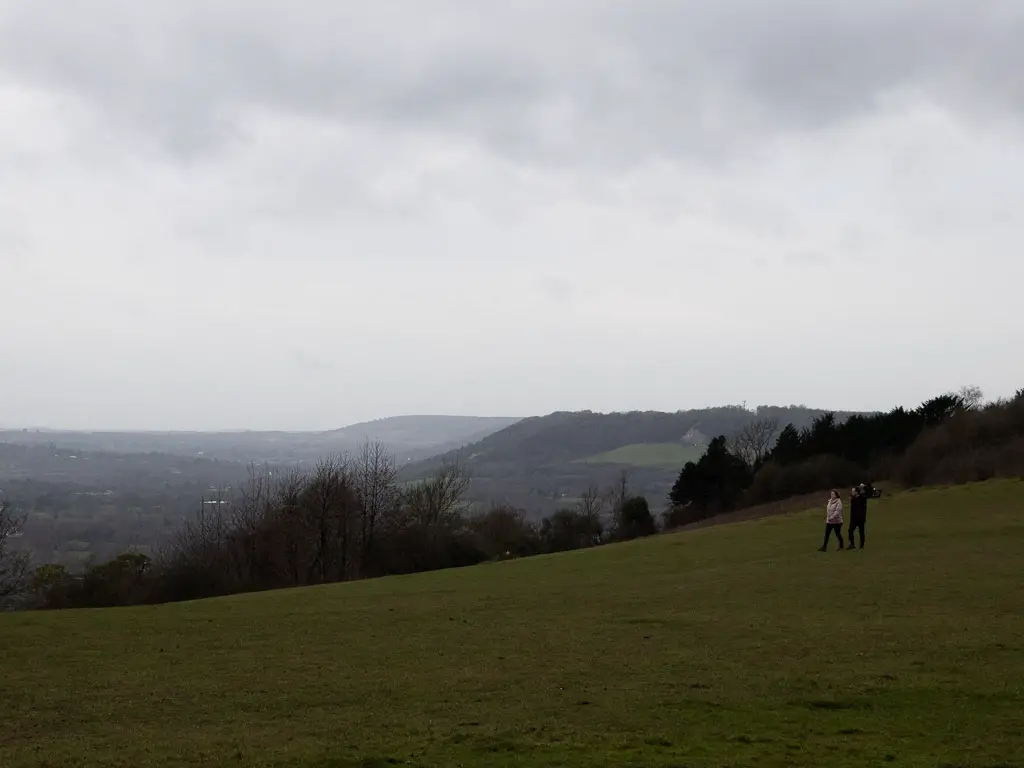 A view across the hilltop to the hills in the distance towards the end of the walk from Westhumble to Merstham along the North Downs Way stage 3 trail. There are two people walking across the hill, and the sky is misty.