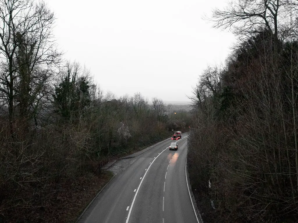 Looking down to an A road on the walk between Westhumble and Merstham along the North Downs Way. The road is lined with leafless trees, and there are a couple of cars on the road.