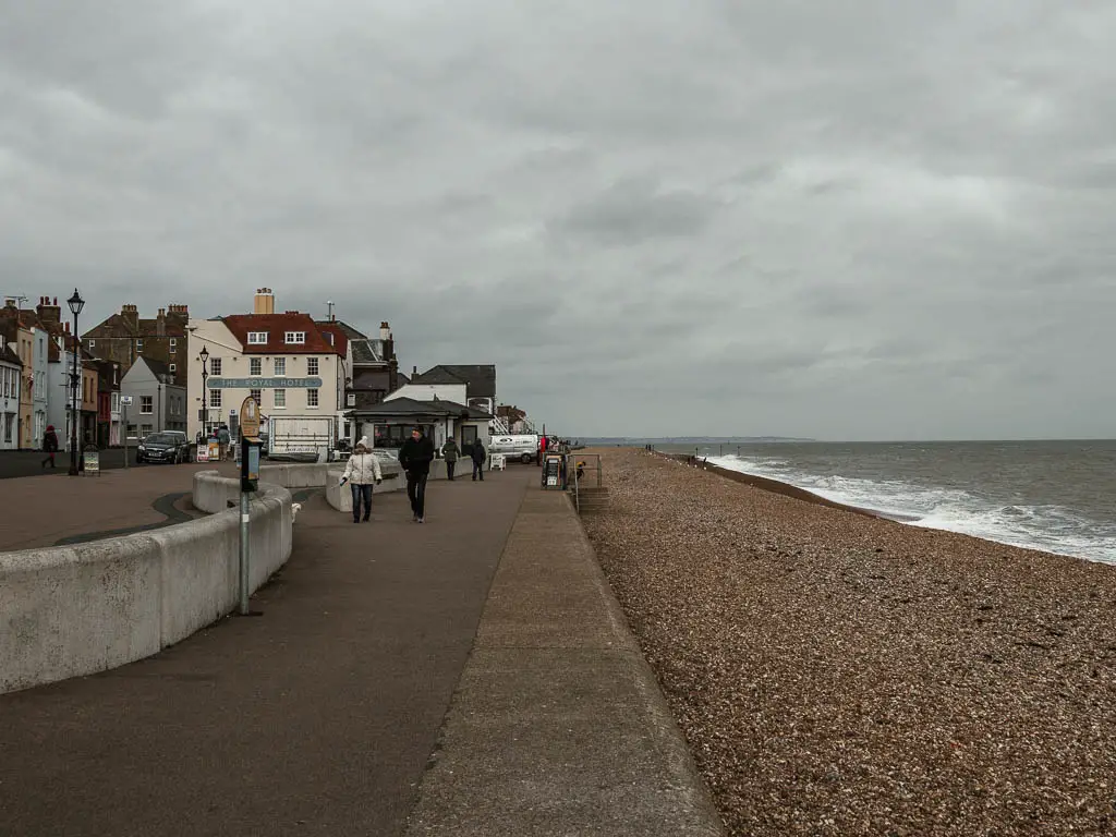 A concrete walkway next to the single beach and English Channel on the walk though Deal. There are a few people walking on the walkway. 